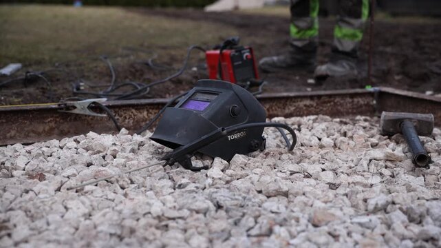 TORROS helmet rests on pale gravel by a rusted rail as leads run to a red unit, a sledgehammer lies nearby, and a worker in high vis trousers stands. Shallow depth of field.