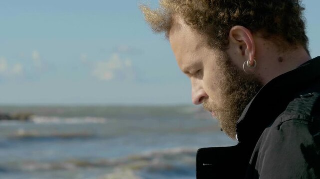 Young brown haired man looking down sadly then raising gaze toward the sea. Emotional outdoor scene expressing reflection, hope and inner thoughts.
