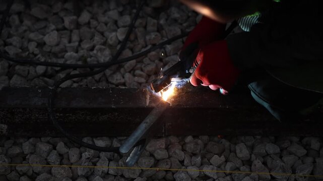 Gloved hands hold welding pliers on a rusted rail as sparks fly and smoke rises over loose ballast, shallow depth of field and low exposure show tool motion.