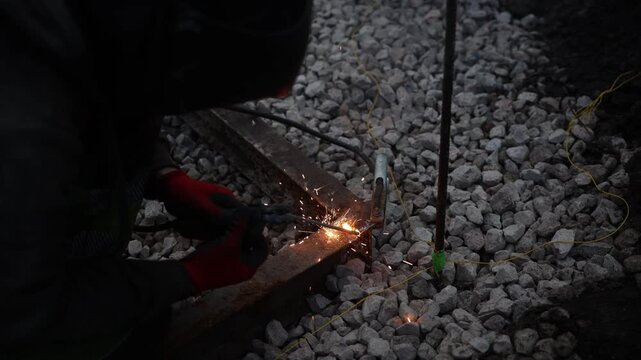 Worker in welding helmet and red gloves joins metal flange to rebar over gravel ballast, bright arc sparks and smoke rise, thin yellow cord and green tape mark rebar
