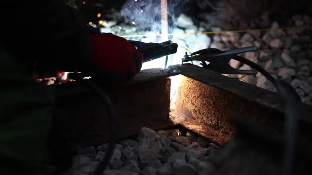 Worker in red glove holds electrode as arc melts rail joint. Sparks and smoke rise, molten seam glows orange. Clamps and cables sit on ballast in low light.