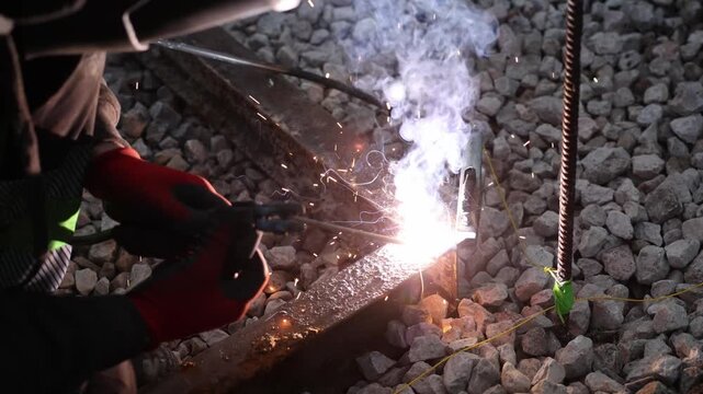 Worker in red gloves manipulates electrode holder as arc emits sparks, smoke, and molten metal on rusted rail over coarse ballast with rebar tied by yellow cord and tape.