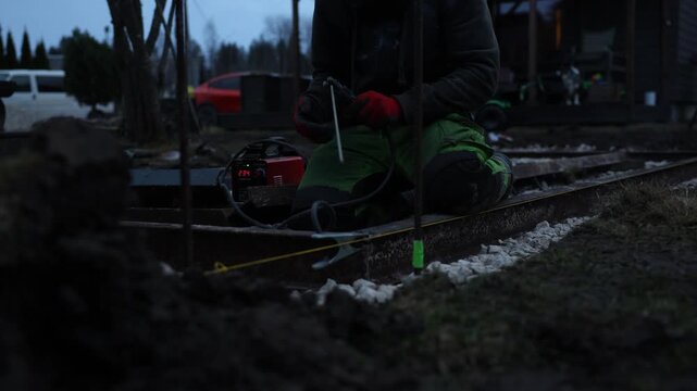 Worker kneels with helmet and red gloves, operates small red welder showing 234, guides rod on rusty rails, yellow string and green taped stake mark alignment.