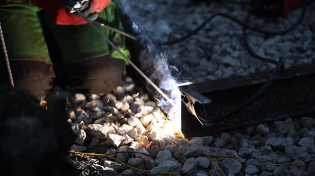 Worker welds steel rail with electrode as sparks and smoke rise, heavy clamp holds joint, thick cable carries current, gravel ballast, boots, gloves, low light, shallow depth