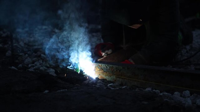 Low light close up shows welder kneeling by rusted rail, arc welding along a string line. White blue arc, orange molten metal, sparks, and smoke rise in night repair.