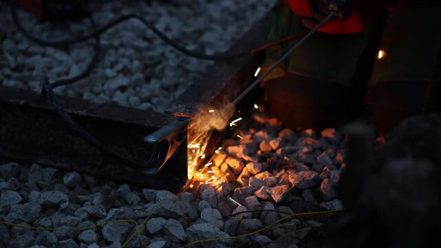Night rail welding with sparks and smoke, worker kneels on ballast. A close view shows glowing metal at a joint, clamps and cables, tight framing, shallow depth of field.