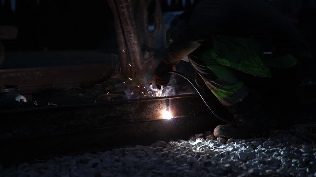 Worker crouches on rail line and welds a seam at night, bright arc casts cool tone, orange sparks fly, smoke drifts, ballast stones and tree trunk sit nearby.
