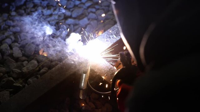 Welder performs rail joining at a track crossing at night, with arc light, flying sparks, molten beads, and smoke over ballast stones, handheld and shallow depth of field.