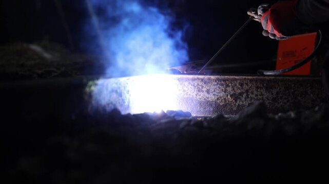 Low angle close up shows a worker welding a rusted rail at night, arc glows, sparks and smoke cross the ballast, portable unit at right, molten seam forms.