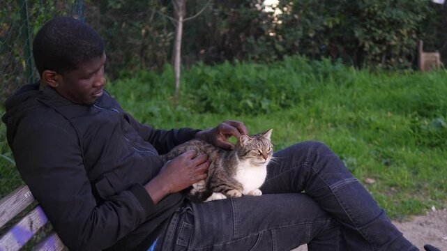 Side view of a young African American man in a black jacket gently petting a cat in the park. The scene captures tenderness, calm, and connection with nature.