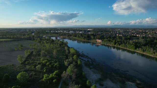 Po River flowing through countryside near Cremona, Lombardy, Italy, with floodplain vegetation, agricultural fields, urban outskirts and distant iron bridge across river, drone panning clockwise