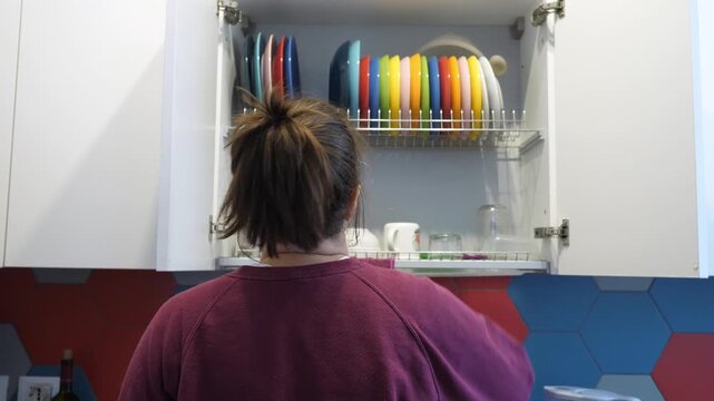 Young woman trying to take an item from dish rack but suddenly feeling neck pain. Realistic kitchen scene showing strain, discomfort and everyday home activity.