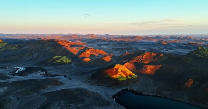 Aerial view of volcanic highlands at golden hour, with orange-lit cones, dark lava plains, winding channels, and a deep blue crater lake in a vast remote landscape.