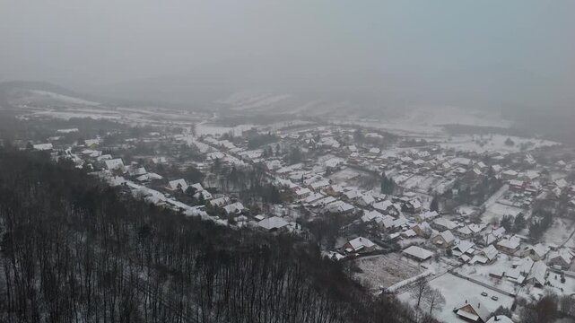 Pilisszentl&aacute;szl&oacute;, a remote mountain village in Hungary's Pilis Mountains, features snow-covered houses and bare winter trees surrounded by forested hills in a peaceful countryside from an aerial view.