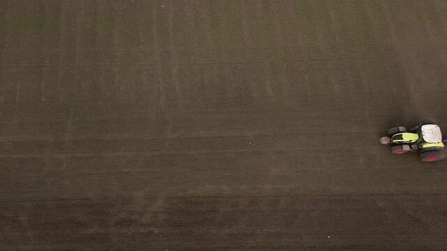 Aerial perspective of a blue tractor working on a brown field, forming furrows in the soil, with a clear sky visible above