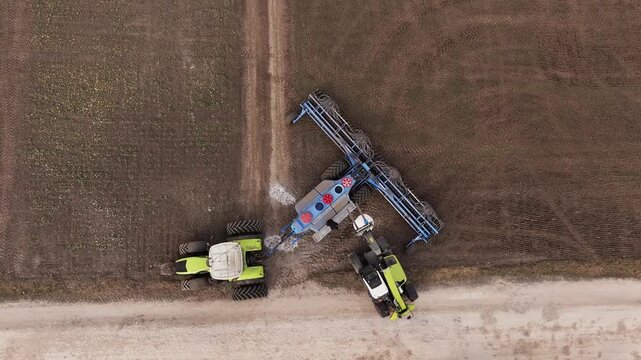 Green telehandler lifts a blue tank while a large plow is attached, set in a farm field with brown soil and green grass in the background