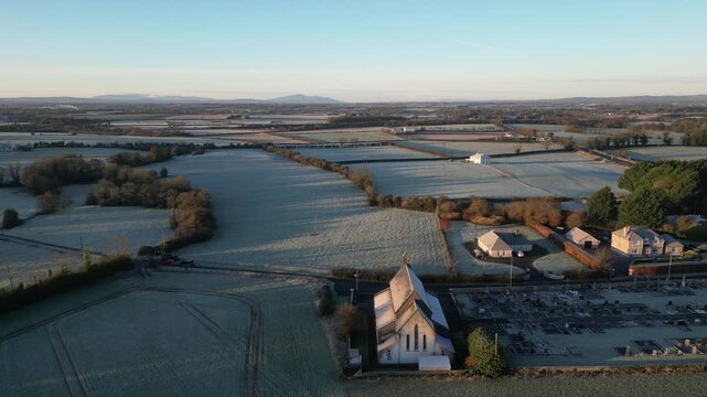 Drone footage captures stunning aerial view of frost-covered Saint Brendan's Church and landscape at dawn, revealing the serene beauty and expanse of the Irish countryside.