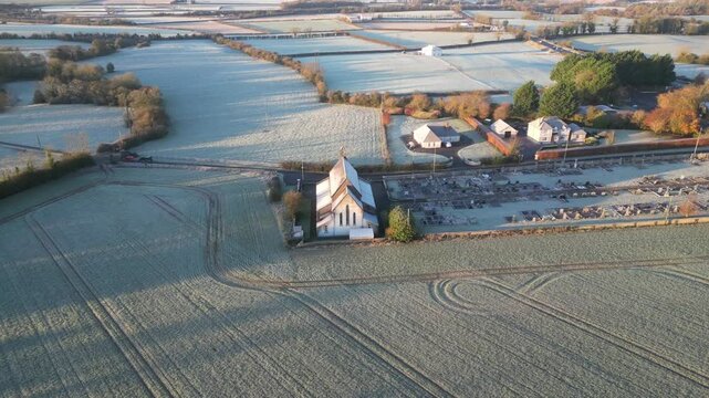 A breathtaking aerial footage showcases Saint Brendan's Church and cemetery surrounded by frost-covered farms in Muckalee, Ireland under the soft, early morning light.