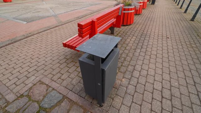 Close-up of a modern public trash bin beside a red bench on cobblestone paving in an empty town square, urban cleanliness and street infrastructure concept.
