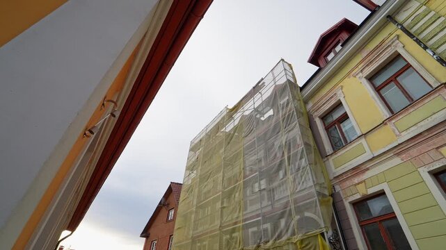 Street view of a historic apartment building under renovation, wrapped scaffolding covered with yellow safety mesh in an old town setting under an overcast sky.