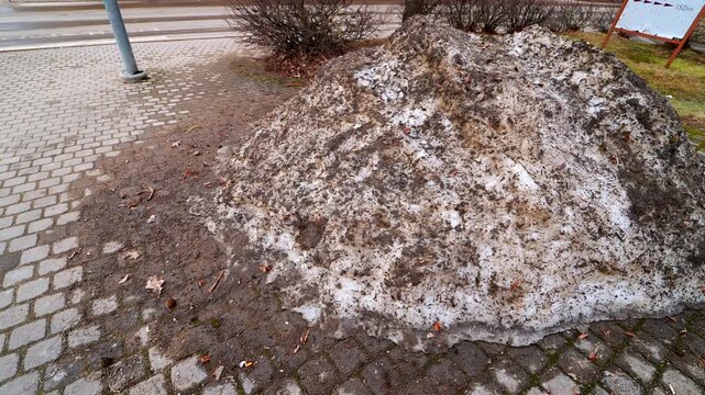 Large dirty snowbank piled beside a city sidewalk after winter, stained with sand and road grime as thawing slush turns into muddy meltwater&mdash;urban spring cleanup and safety concept.