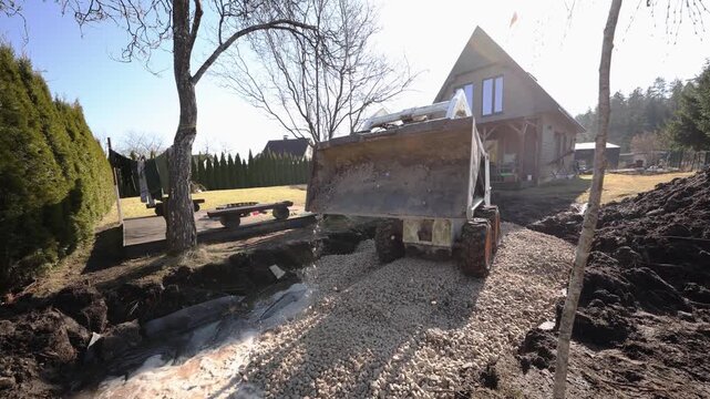 Heavy equipment loads crushed stone into a black lined pond near a triangular roof cottage and wooden deck, clothesline visible, operator maneuvers bucket over gravel path.