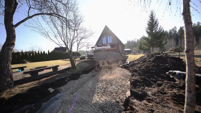 Loader pours gravel into pond liner by triangular roof cabin. Title Loader pours gravel into pond liner by triangular roof cabin. Stones fall in bright midmorning light.