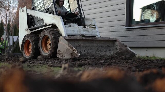 White skid steer with orange rims scrapes soil in a yard, operator drives with controlled motion, muddy tires turn clods near siding and a small ride on tractor.