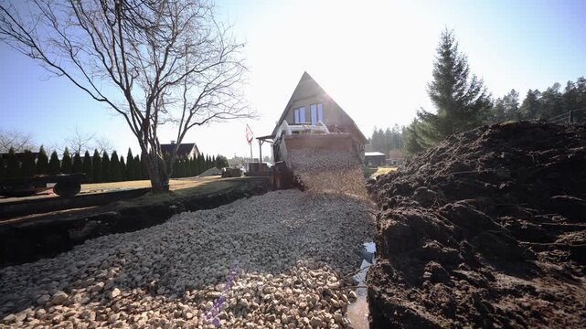 Front end loader empties crushed stone into deep trench by A frame house with flagpole and tall hedge, soil piles on sides, stone dust and runoff in daylight backlight