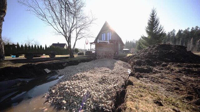 Wide angle, low angle view shows excavator dumping crushed stone into a long trench by a triangular roof house, with water on liner, soil mound, fence, and lens flare.