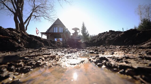 Low angle view shows muddy trench with reflective puddles, excavator by a triangular roof cabin, striped flag, bare trees, dark soil piles, slow excavation motion, title