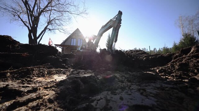 Small turquoise excavator digs soil before triangular roof wooden house. Low angle shows churned mud, lens flare, and operator silhouette in late morning.