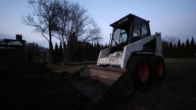 Small white skid steer with metal grid cab and orange wheels sits on muddy ground in a residential yard at early evening, bucket partly filled, leafless tree and conifer hedge present.
