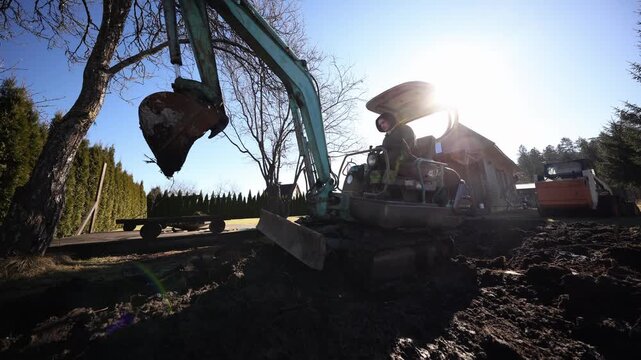 Teal mini excavator with rusted bucket sits on churned mud as operator maneuvers controls beside a steep roof wooden cabin, with hedges, bare tree, and roller.