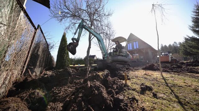 Compact excavator lifts and swings bucket, roots and dark loam churn near chain link fence and steep roof timber house, roller compactor parked, bright midday wide angle