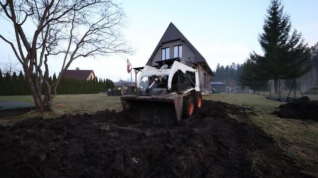 Skid steer with rusted bucket pushes churned soil near steep roof house. Operator sits in cab. Leafless tree left, tall conifer right, wide angle, cold light.
