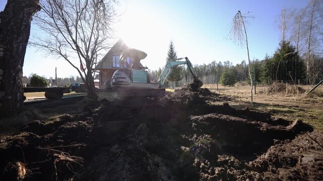 Small turquoise excavator digs soil by triangular roof wooden house. Low angle wide frame shows root filled earth, birch trunks, wire fence, lens flare, and active arm.