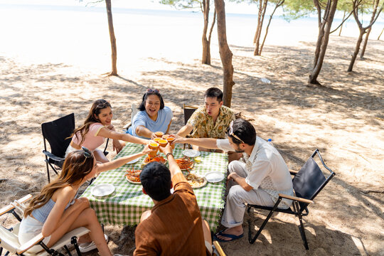 Group of Happy Asian friends toasting drinks and sharing seafood meal together at outdoor picnic table, people enjoying summer beach holiday vacation active lifestyle social gathering party concept