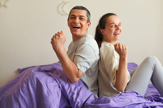 A man and woman sit on a bed with purple bedding. They face opposite directions but share laughter, celebrating their morning together