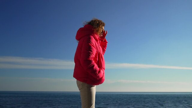 Seaside woman leisure young female wearing a bright pink puffer jacket standing by the ocean under a clear blue sky