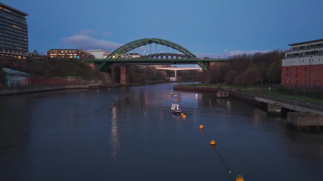 Aerial drone shot flying forward and climbing over the River Wear in Sunderland at dawn, revealing the illuminated Wearmouth Bridge and Monkwearmouth Railway Bridge.