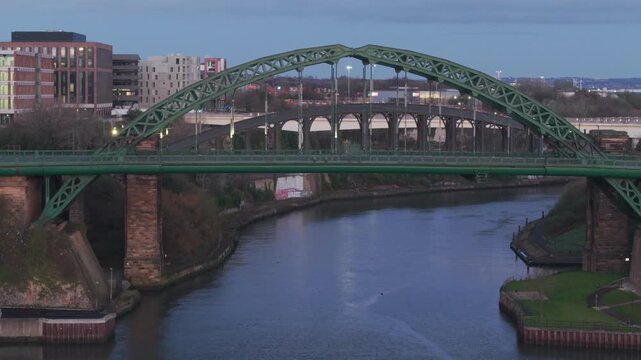 Aerial telephoto tracking shot of the Wearmouth Bridge over the River Wear at dawn, with city lights still visible and reflecting on the water.