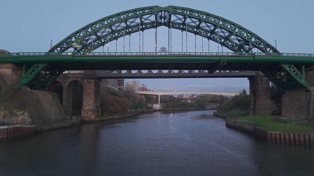 Aerial drone shot flying slowly under the Wearmouth Bridge and Monkwearmouth Railway Bridge at dawn, with the River Wear below and the modern Keel Crossing visible in the far background.