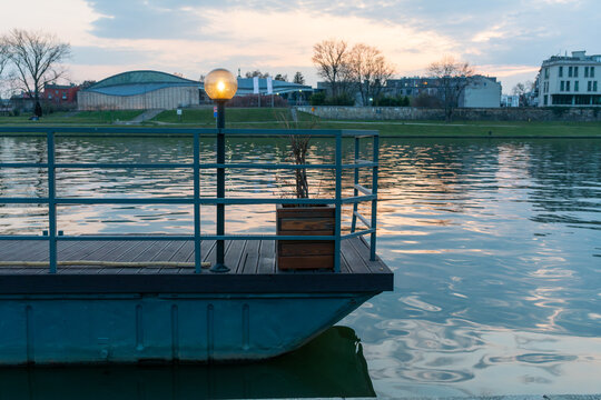 Metal floating deck with wooden floor, railing, glowing lamp, and planter set against calm evening water. Soft sunset light and shoreline architecture create a peaceful waterfront, mooring, and rivers
