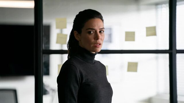Serious businesswoman standing by glass wall with sticky notes in modern office, focused professional reflecting on planning, strategy and workplace decision making