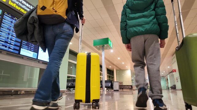 Family walking with luggage in an airport terminal, the boy pulling a green suitcase and the mother a yellow one, viewed from behind at a low angle while moving forward