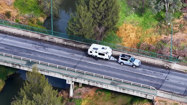 Aerial view of white suv towing caravan and large livestock truck crossing rural bridge over river.