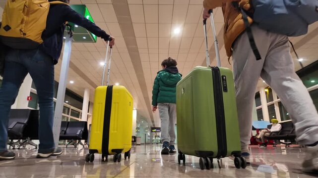 Family with child walking through the airport terminal with luggage, pulling colorful suitcases toward the boarding gate for their vacation journey. Low-angle view of travel