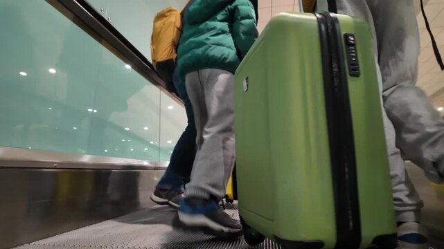 Family with child and luggage using moving walkway at the airport terminal. Rear view of travelers with backpacks and suitcases going to their flight departure gate