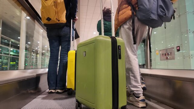 People with luggage and backpacks using moving walkway at airport terminal. Back view of unrecognizable passengers with suitcases traveling on a departure terminal autowalk conveyor belt